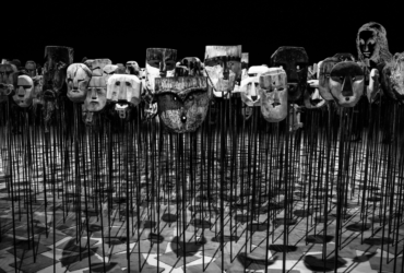 A black & white Image of a variety of wooden masks atop vertical sticks with a sketch of the mona lisa amongst them depicting the mask of mona lisa.